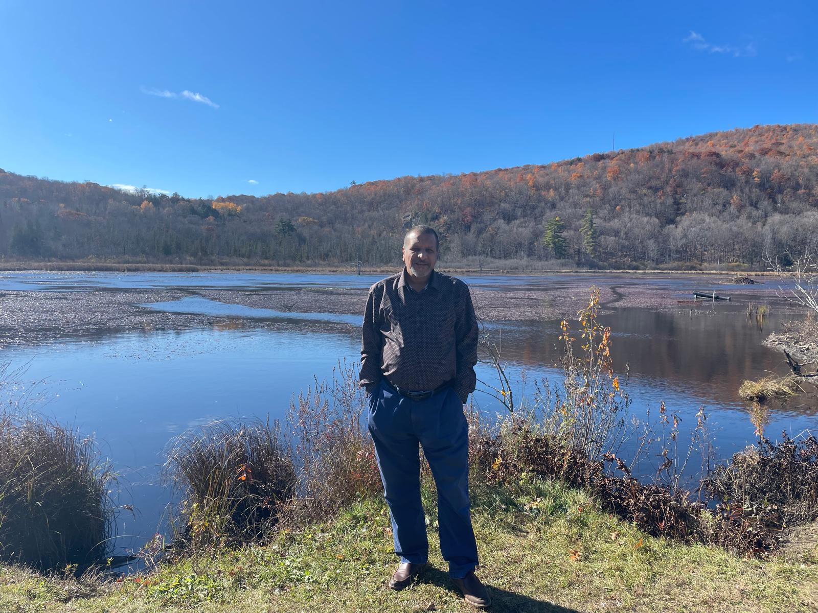 Adam Zahy standing outdoors near a calm lake, with grassy plants in the foreground, forested hills in the background, and a clear blue sky above. The scene appears peaceful and natural, with autumn colours visible on the trees.