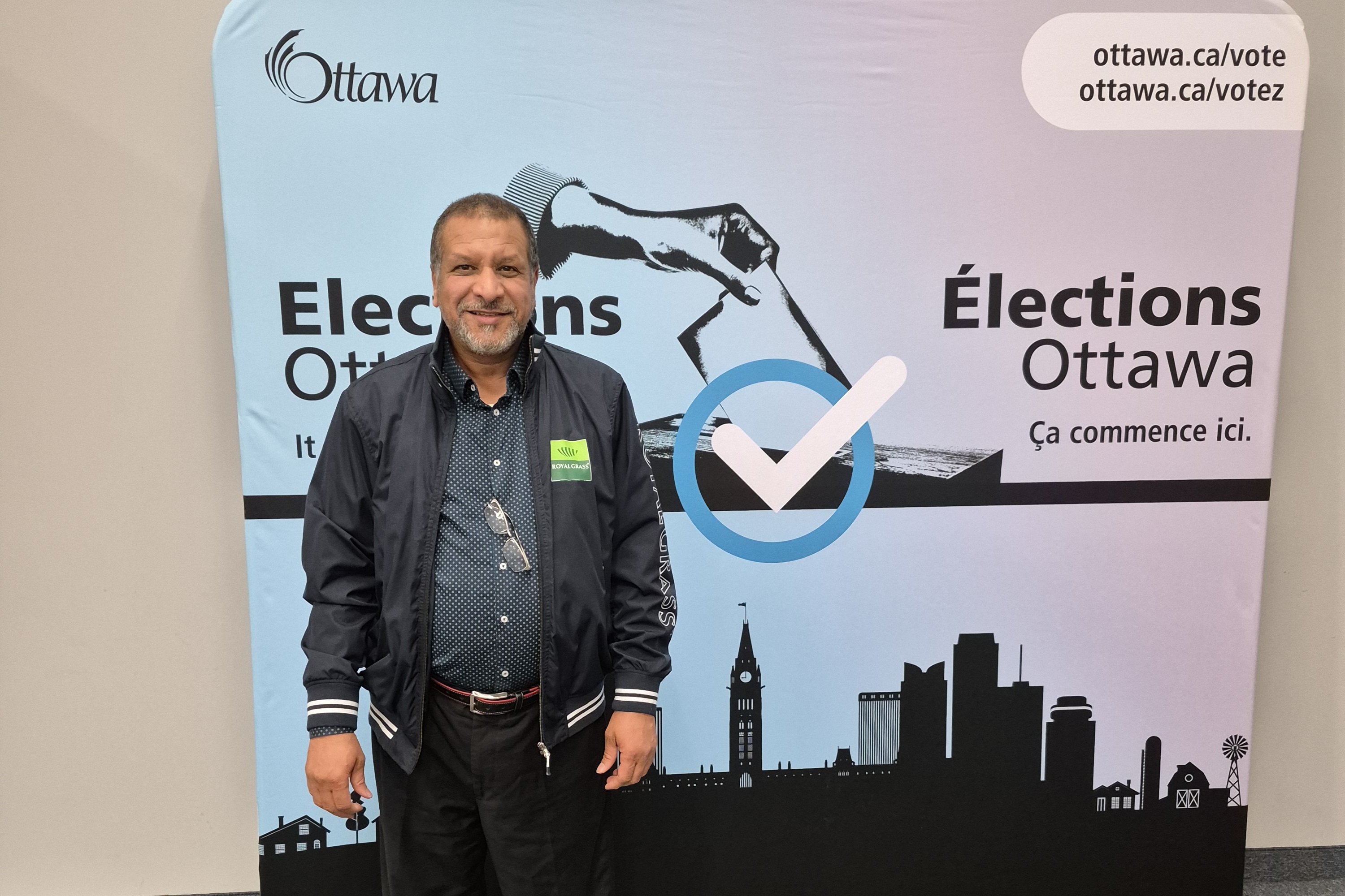 Adam Zahy standing in front of an Elections Ottawa display with a large checkmark graphic and Ottawa skyline silhouette. The display includes English and French text reading “Elections Ottawa” and “Élections Ottawa.”